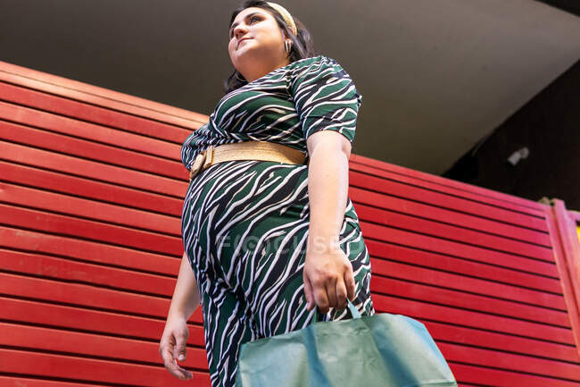 Vista lateral de ángulo bajo de la joven mujer con curvas felices en vestido elegante con impresión geométrica que lleva bolsas de compras mientras está de pie cerca de la pared de rayas rojas en la ciudad - foto de stock