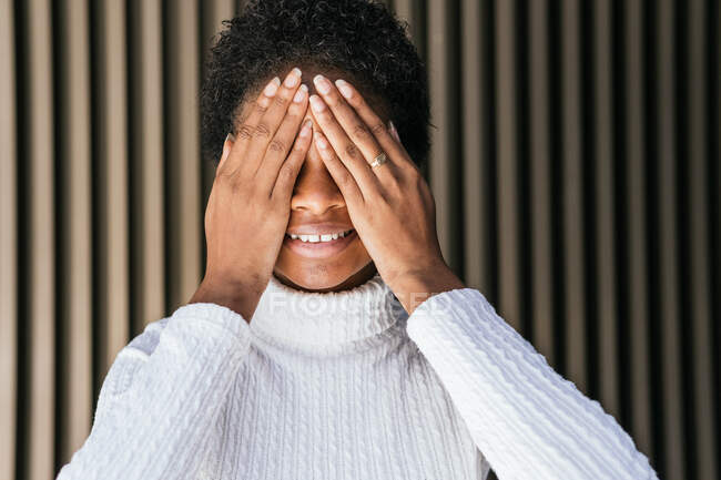 Mulher afro-americana feliz em camisola na moda cobrindo os olhos com sorriso contra a parede listrada na rua — Fotografia de Stock