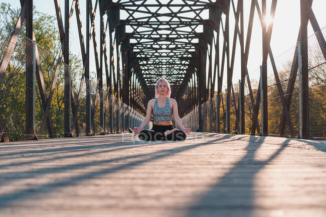 Ground level of female with closed eyes sitting in Padmasana pose while practicing yoga on bridge footpath in sunlight — Stock Photo