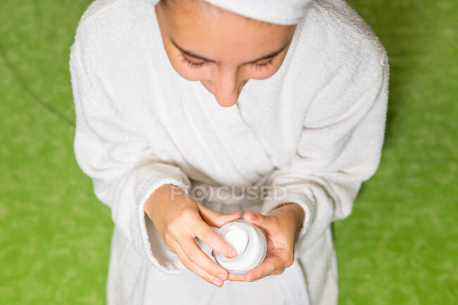 Visão superior da jovem fêmea em roupão de banho branco sentado na cama e tomando creme de jarra durante a rotina de cuidados com a pele em casa — Fotografia de Stock