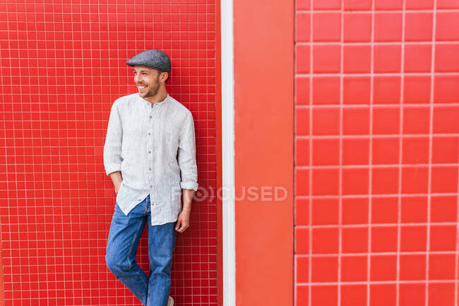Guapo joven barbudo en camisa casual de moda y jeans y gorra de pie cerca de la pared roja y mirando hacia otro lado con sonrisa - foto de stock