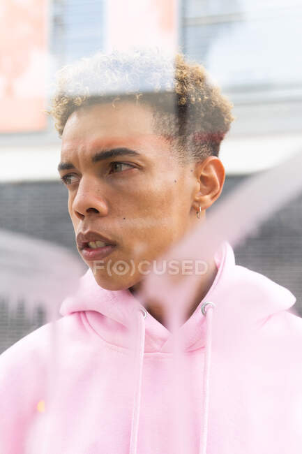 Through glass of serious young ethnic hipster guy with Afro hairstyle dressed in pink hoodie at window and looking away — Stock Photo