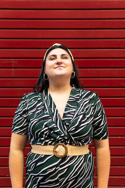 Portrait of cheerful young curvy brunette in stylish striped outfit and headband while have closed eyes against red wall — Stock Photo