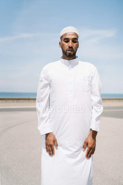 Full boy Islamic male in traditional white clothes standing on rug and praying against blue sky on beach — Stock Photo