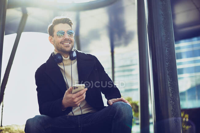 Contenido masculino en ropa elegante con auriculares y teléfono celular sentado en la ciudad en el día soleado - foto de stock