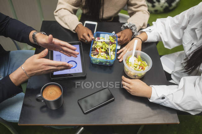 Desde arriba de los compañeros de trabajo multirraciales irreconocibles recortados sentados a la mesa y almorzando durante el descanso en la oficina - foto de stock