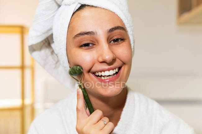 Giovane donna felice con asciugamano sulla testa guardando la fotocamera con sorriso e massaggio viso con rullo di giada durante la routine di cura della pelle a casa — Foto stock