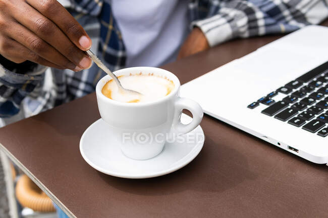 Crop anônimo étnico masculino freelancer mexendo café quente na xícara colocada na mesa com laptop no café ao ar livre — Fotografia de Stock