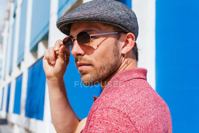 Young bearded guy wearing casual red polo shirt and cap standing against blue wall on street in summer day — Stock Photo