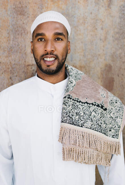 Homme islamique dans des vêtements blancs authentiques tout en se tenant contre le mur grogneux — Photo de stock
