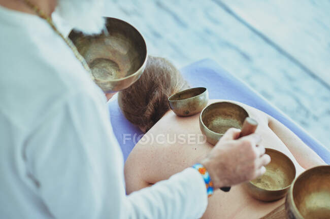 From above of crop unrecognizable male spiritual practitioner playing Himalayan bowls on back of topless female — Stock Photo