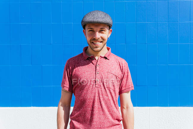 Радісний молодий бородатий чоловік у стильному повсякденному одязі та кепці насолоджується літнім днем біля синьої стіни на міській вулиці дивлячись на камеру — стокове фото