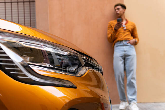 Молодой стильный этнический кудрявый парень в модном наряде, используя смартфон, прислонившись к стене рядом с припаркованным современным оранжевым автомобилем на городской улице — стоковое фото