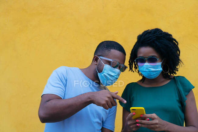 Coppia nera con maschera utilizzando un telefono cellulare appoggiato a una parete gialla — Foto stock