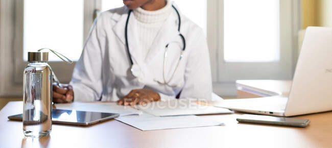 Crop médico feminino preto irreconhecível com estetoscópio escrevendo informações na folha de papel enquanto prepara o relatório médico à mesa no escritório da clínica moderna — Fotografia de Stock