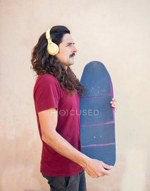 Vue latérale du skateboarder masculin avec des cheveux ondulés écoutant la chanson des écouteurs sans fil tout en regardant vers l'avant sur fond beige — Photo de stock