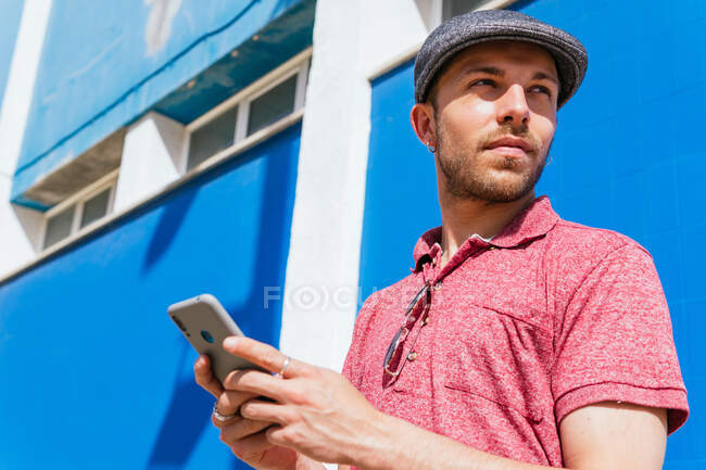 Снизу задумчивый молодой бородатый хипстер в повседневной рубашке для поло и кепке просматривает мобильный телефон, стоя напротив синей стены в солнечном свете, отворачиваясь — стоковое фото