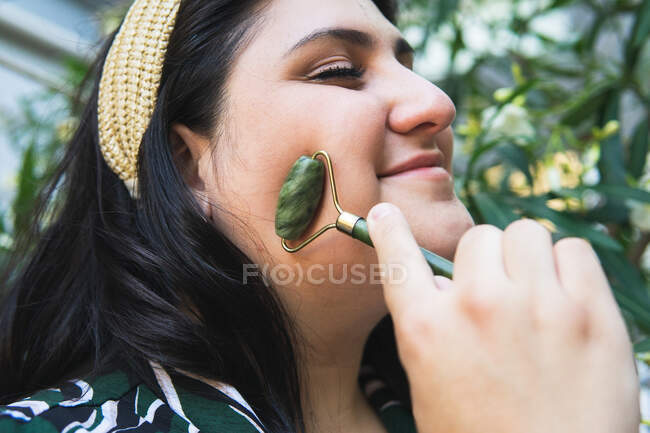 Seitenansicht positiver junger kurviger Frauen, die Gesichtsmassage mit grüner Jadewalze während der Hautpflege genießen — Stockfoto