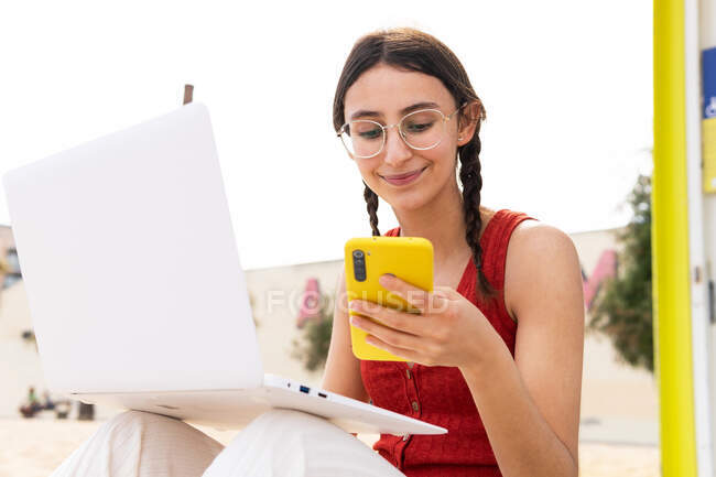 Contenu femme pigiste assise sur le bord de la mer et tapant sur ordinateur portable et naviguant sur smartphone tout en travaillant à distance sur le projet en été — Photo de stock