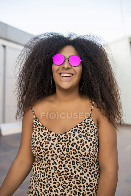 Mujer joven y afro con gafas de sol riendo y sonriendo mientras mira a la cámara - foto de stock