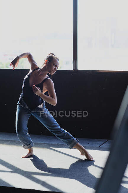 Jeune femme pieds nus en jeans avec un chignon de cheveux dansant tout en regardant vers le bas sur le sol avec des ombres au soleil — Photo de stock
