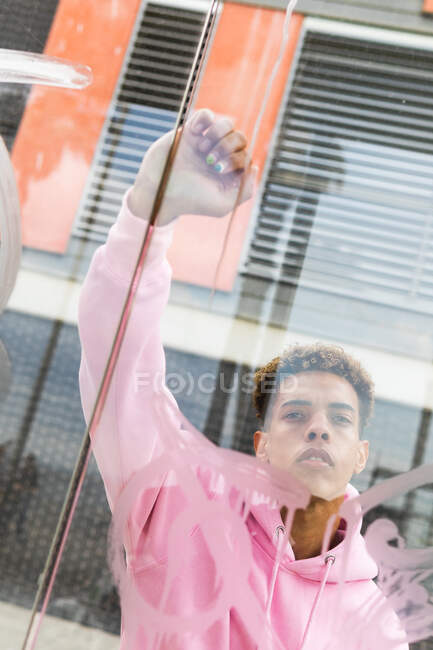 Through glass of serious young ethnic hipster guy with Afro hairstyle and manicure dressed in pink hoodie knocking at window and looking at camera — Stock Photo