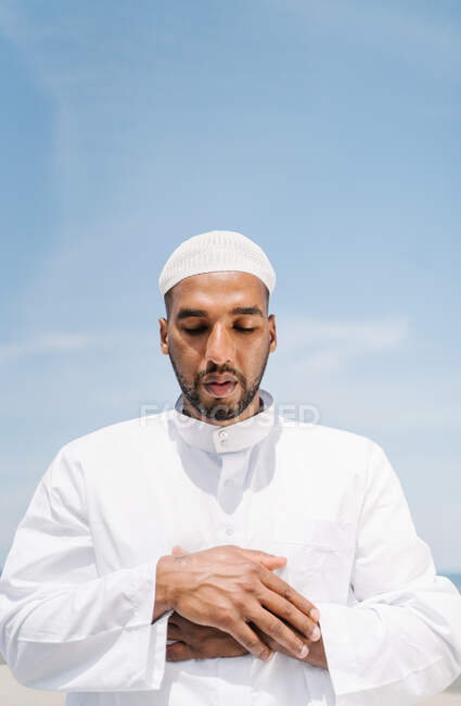 Menino completo macho islâmico em roupas brancas tradicionais em pé no tapete e orando contra o céu azul na praia — Fotografia de Stock
