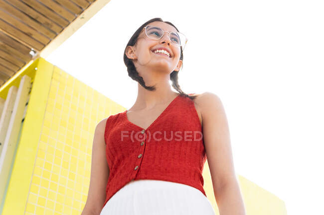 Low angle of young female in stylish glasses and with pigtails standing on background of bridge in city and looking away — Stock Photo