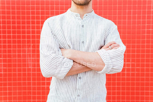 Неузнаваемый бородатый самец в модной полосатой рубашке, стоящей с руками, скрещенными к красной стене — стоковое фото