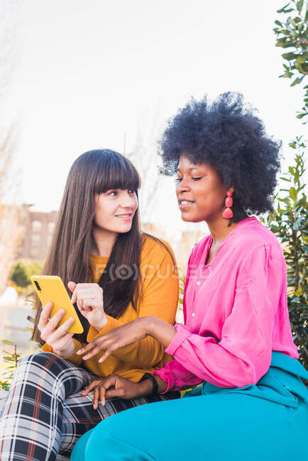 Feliz pareja multiétnica de mujeres homosexuales sentadas en la calle y navegando juntos por el teléfono móvil - foto de stock