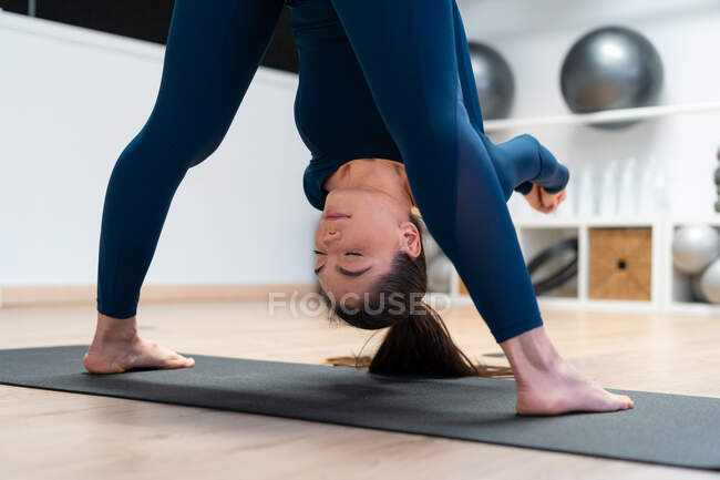 Vista posterior de la joven hembra flexible enfocada haciendo pose Standing Forward Bend with Shoulder Opener mientras practica yoga en un gimnasio - foto de stock