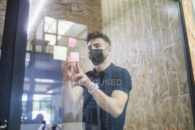 Мужчина исполнительный директор пишет на липкой ноте на стеклянной стене во время мозгового штурма с коллегами в офисе — стоковое фото
