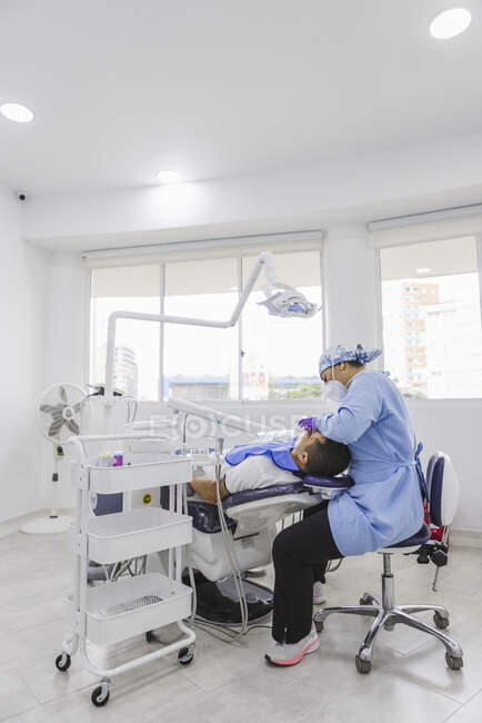 Vista lateral médica femenina en uniforme de limpieza de dientes de paciente masculino con retractor dental en el hospital - foto de stock