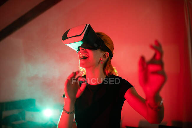 Mulher feliz vestindo t-shirt ao usar óculos VR e de pé em estúdio com luzes de néon vermelho — Fotografia de Stock