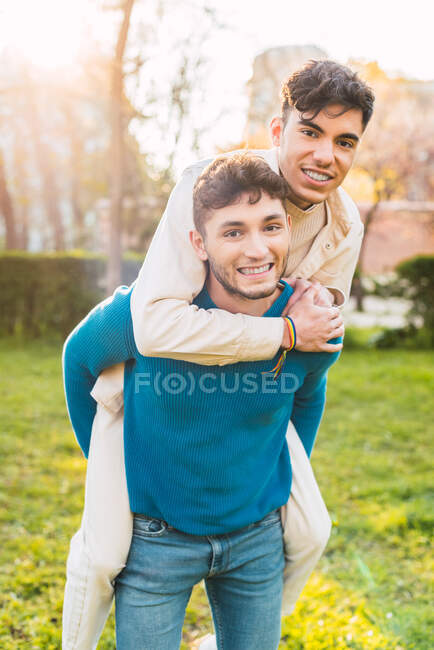 Positivo gay masculino piggybacking sorrindo namorado enquanto se divertindo juntos no parque no fim de semana — Fotografia de Stock