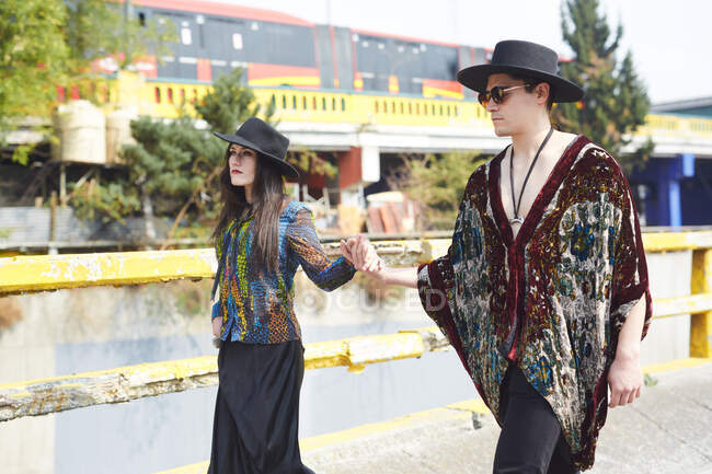 Решительная стильная пара в хипстерской одежде, держащаяся за руки и идущая по улице летом — стоковое фото