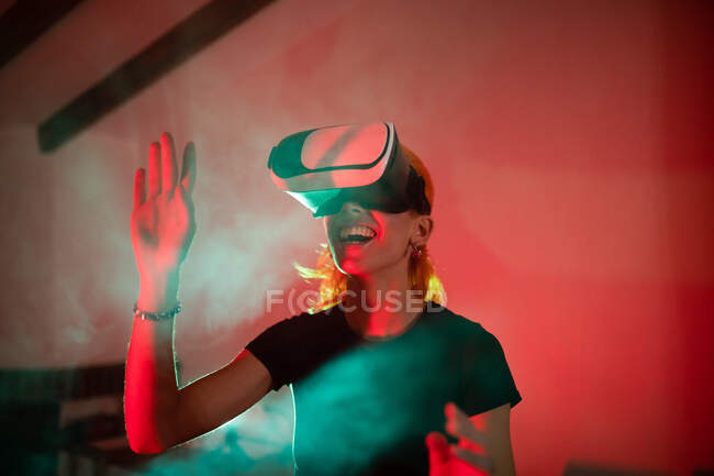 Mulher feliz vestindo t-shirt ao usar óculos VR e de pé em estúdio com luzes de néon vermelho — Fotografia de Stock