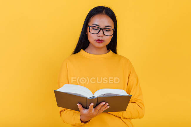 Estudante asiática inteligente em óculos lendo o livro didático e se preparando para o exame em fundo amarelo no estúdio — Fotografia de Stock