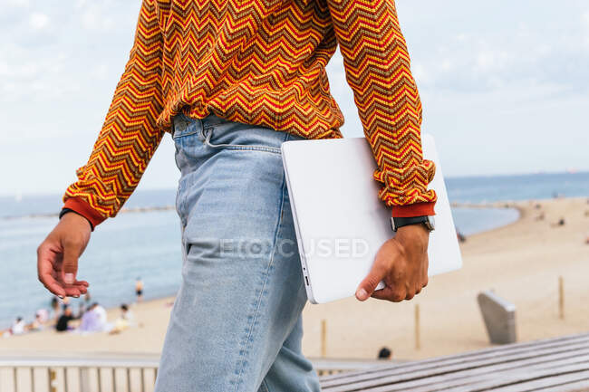 Side view of crop anonymous ethnic male in stylish colorful striped shirt and jeans carrying laptop while walking on embankment near sandy beach and sea — Stock Photo