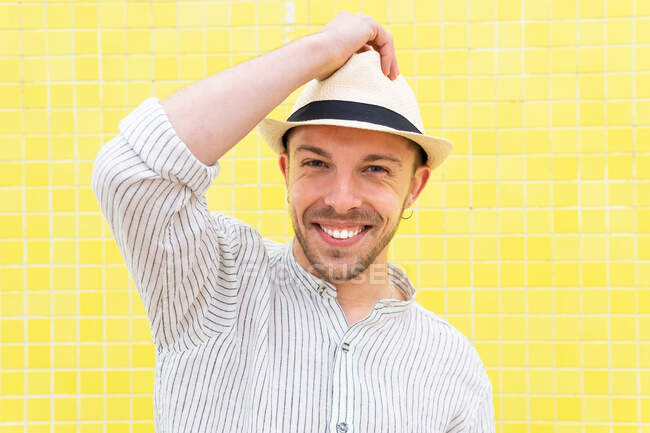 Feliz jovem barbudo hipster cara na roupa da moda e chapéu sorrindo olhando para a câmera enquanto refrigerando sozinho contra a parede amarela no dia de verão — Fotografia de Stock
