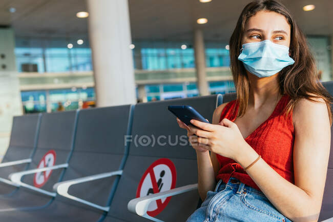 Жінка-туристка в захисній масці сидить у від'їзді в аеропорту і чекає польоту під час епідемії коронавірусу під час використання смартфона — стокове фото