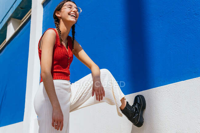 From below of positive female in trendy clothes leaning on wall of building and laughing on sunny day in city street — Stock Photo