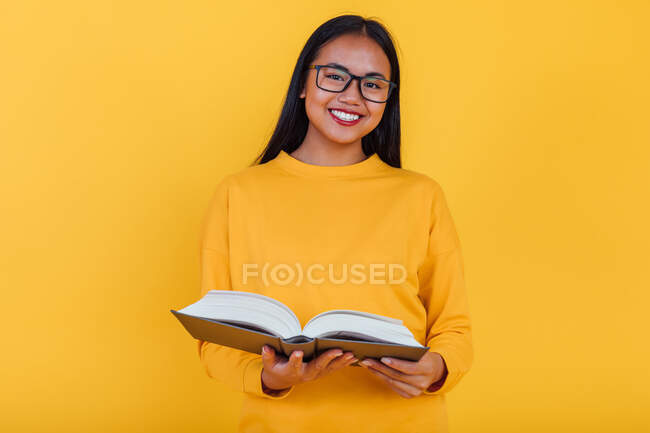 Clever cheerful Asian female student in glasses reading textbook and preparing for exam on yellow background in studio looking at camera — Stock Photo