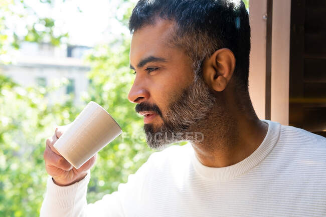 Врожай чоловічий бородатий чоловік п'є каву з керамічної чашки на балконі в сонячний день — стокове фото