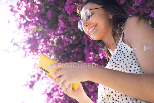 Знизу молода весела жінка використовує телефон у сонячний день у парку з фіолетовими квітами на фоні — стокове фото