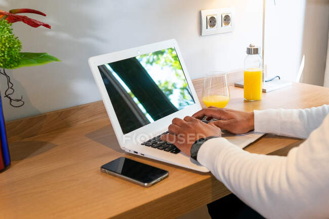High angle of crop anonymous ethnic businessman typing on netbook sitting at table in hotel room during business trip — Stock Photo