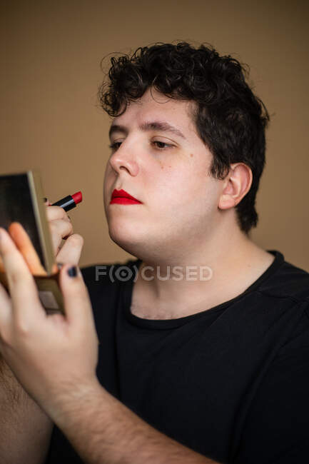 Hombre andrógino femenino con pelo rizado que aplica lápiz labial en los labios mientras hace maquillaje - foto de stock