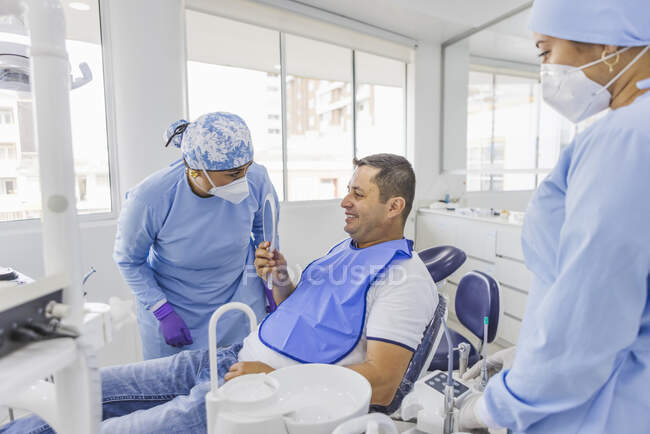 Smiling man looking in mirror while talking to female doctors in uniforms after dental procedure in clinic — Stock Photo