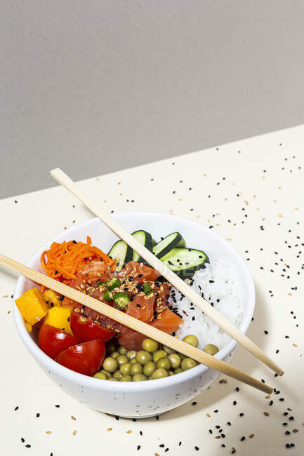 De dessus baguettes de bambou placé sur le dessus du bol avec plat poke savoureux sur la table couverte de graines de sésame — Photo de stock