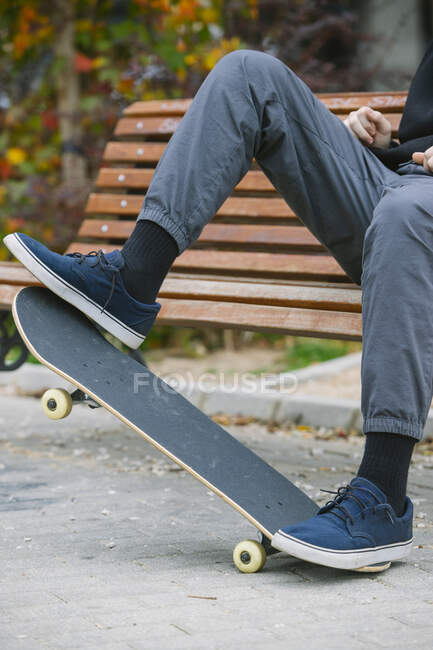 Homme méconnaissable en baskets équilibrant sur skateboard tout en étant assis sur un banc en bois dans le parc avec des buissons pendant l'entraînement en ville — Photo de stock